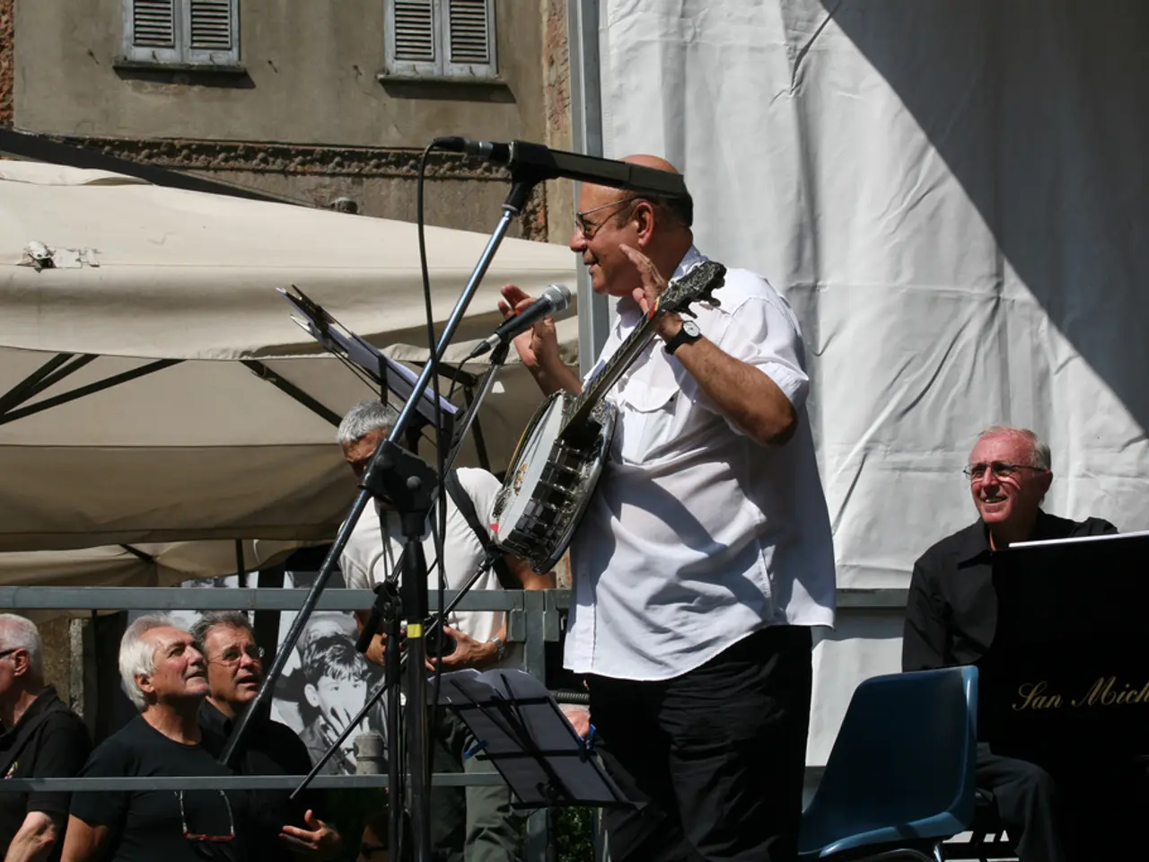 There is an old man talking on mic he is carrying a banjo. in front of him there is stand on it...